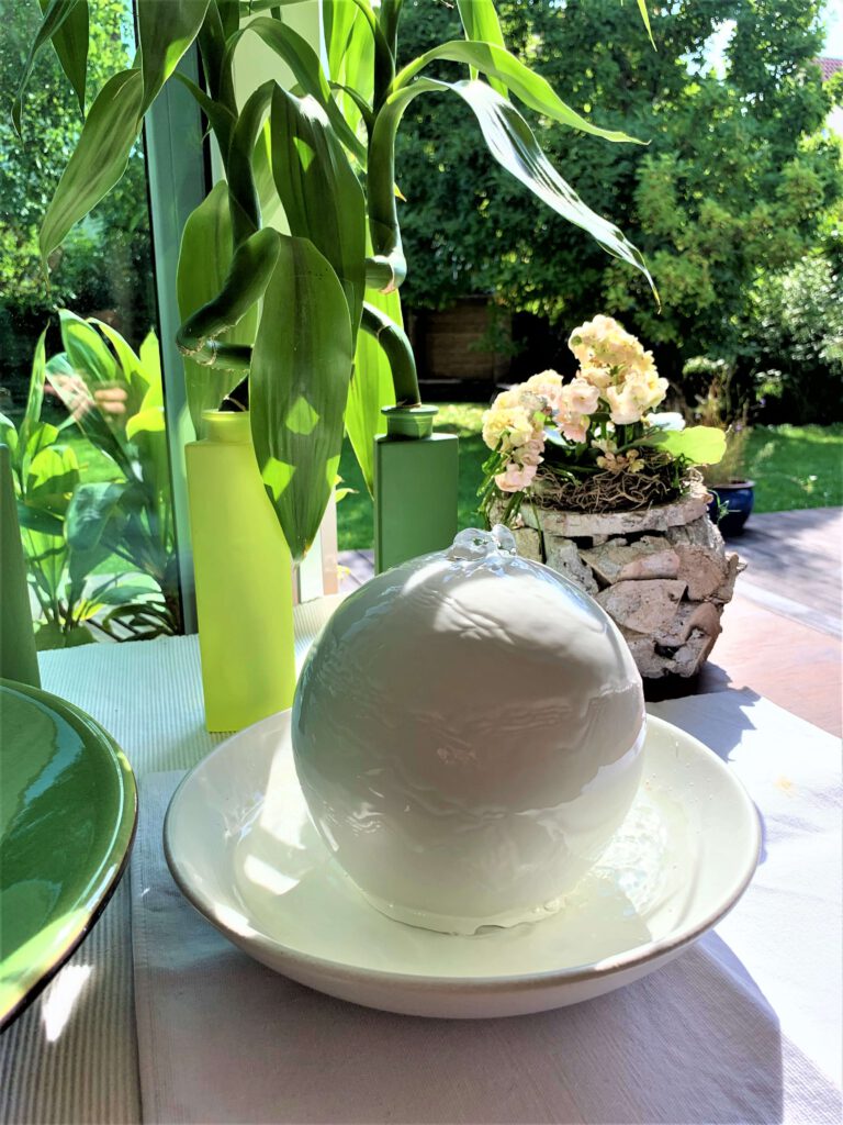 Weißer Kugelzimmerbrunnen steht vor grünem Bambus und Zimmerrose mit Blick in den Garten. Ein sehr vitales, helles, lichtdurchflutetes, energiereiches Bild.